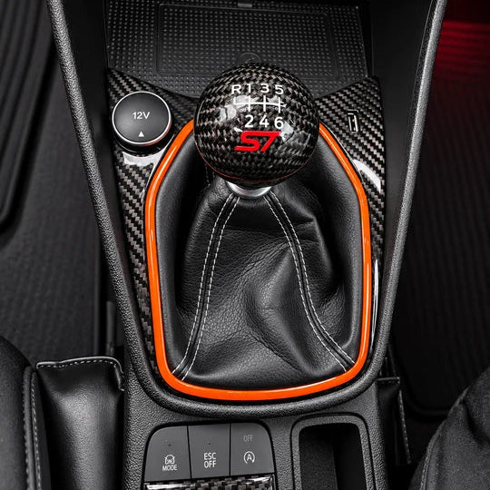 Close-up of a car center console with a black leather gear shift boot, carbon fiber trim, and bright orange accents around the manual gear selector and console.