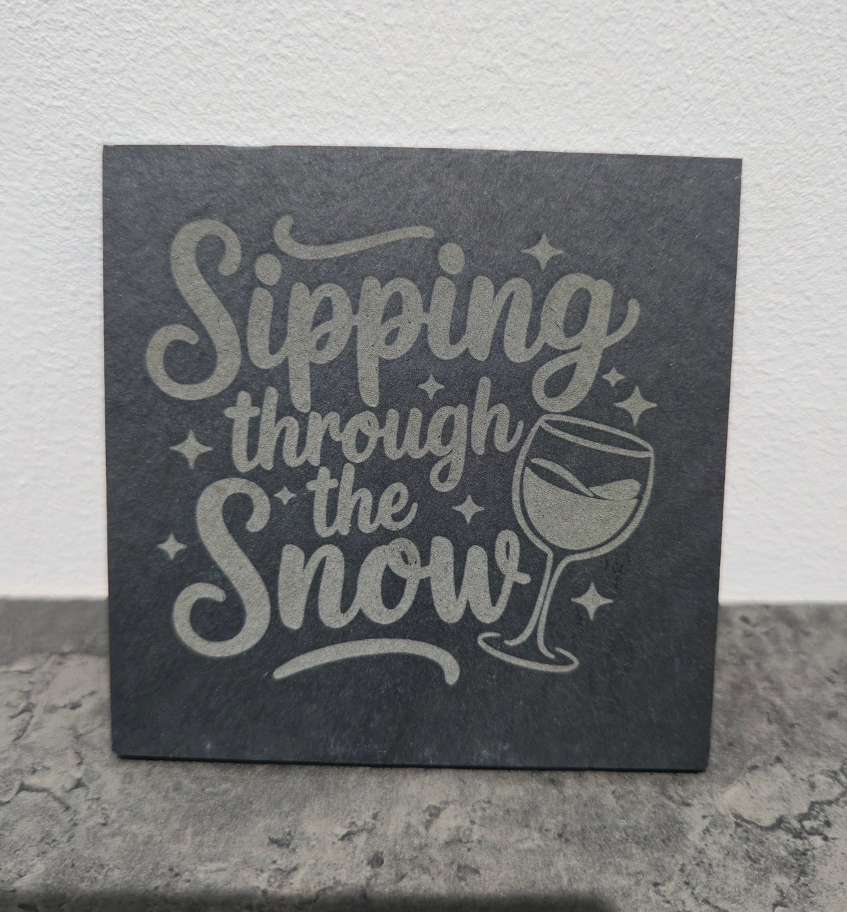 Slate coaster with 'Sipping through the Snow' text and wine glass illustration on a stone surface.