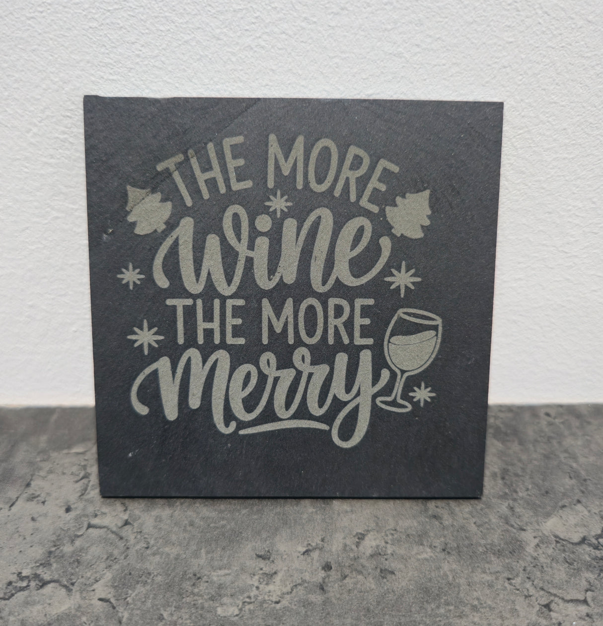 Slate coaster with 'The More Wine The More Merry' text and wine glass illustration on a gray surface.
