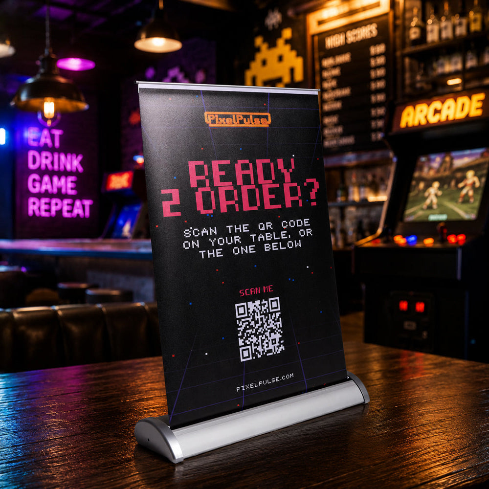 A freestanding black sign on a bar table in a dimly lit pub, with neon signs and an arcade machine blurred in the background. The sign reads “READY 2 ORDER?” with a QR code and small logo text at the top.