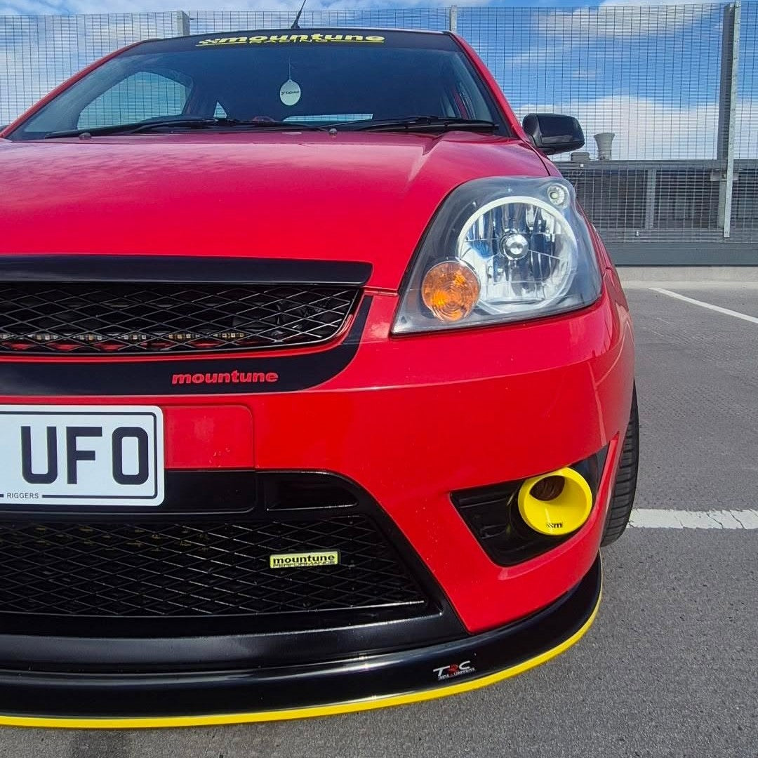 A red car front bumper and lower grille area shown outdoors on a paved surface, with visible yellow trim accents and black grille details. The word 'mountune' appears on the grille and lower bumper, and a license plate reading 'UFO' is visible.
