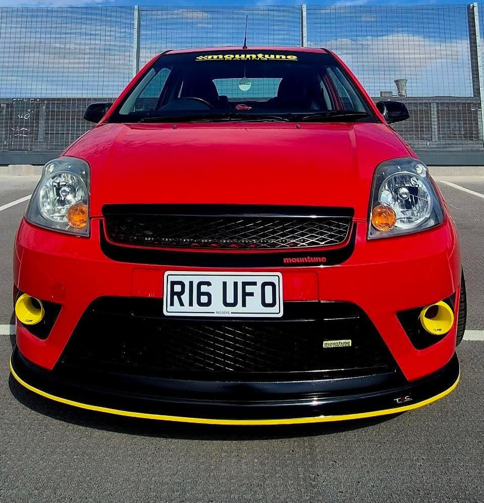 A red sports car parked outdoors on asphalt in front of a fence, with a prominent black front lip spoiler featuring a yellow stripe and matching yellow accents around the fog light openings.