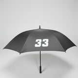 A gray umbrella with the white number 33 printed on the canopy, photographed indoors on a dark carpeted floor with office furniture partially visible at the edges.