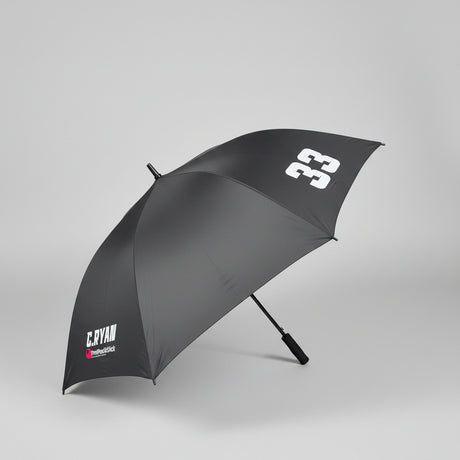 A dark gray golf umbrella with white printed logos, shown open on a gray carpeted floor in an office setting.