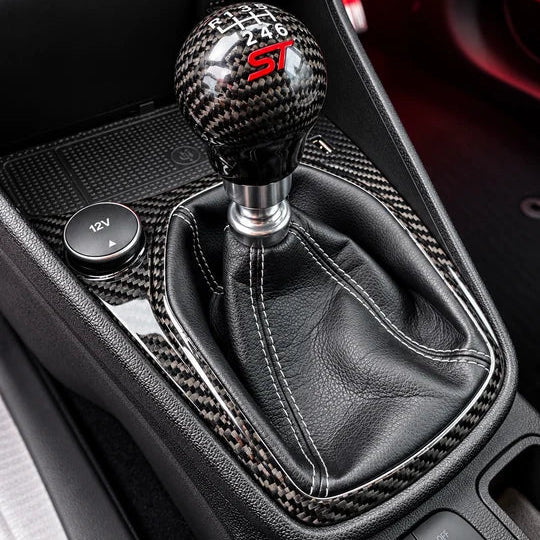 Close-up of a car's manual gear shift knob and boot. The shift knob has a black carbon fiber pattern with a red 'S1' logo, and the boot is covered in black leather with white contrast stitching.