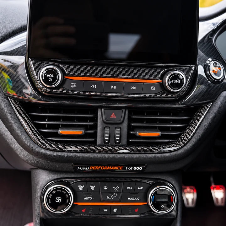 Close-up of a car interior dashboard with carbon fiber trim, a large touchscreen display, climate control buttons, and Ford Performance branding on a black center console.