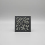 Decorative sign with text 'Surviving Christmas one sip at a time' on a gray background