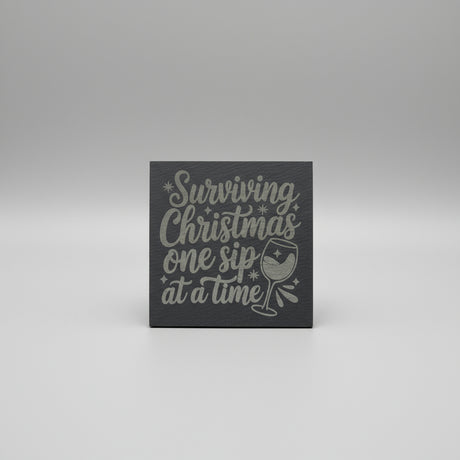 Decorative sign with text 'Surviving Christmas one sip at a time' on a gray background