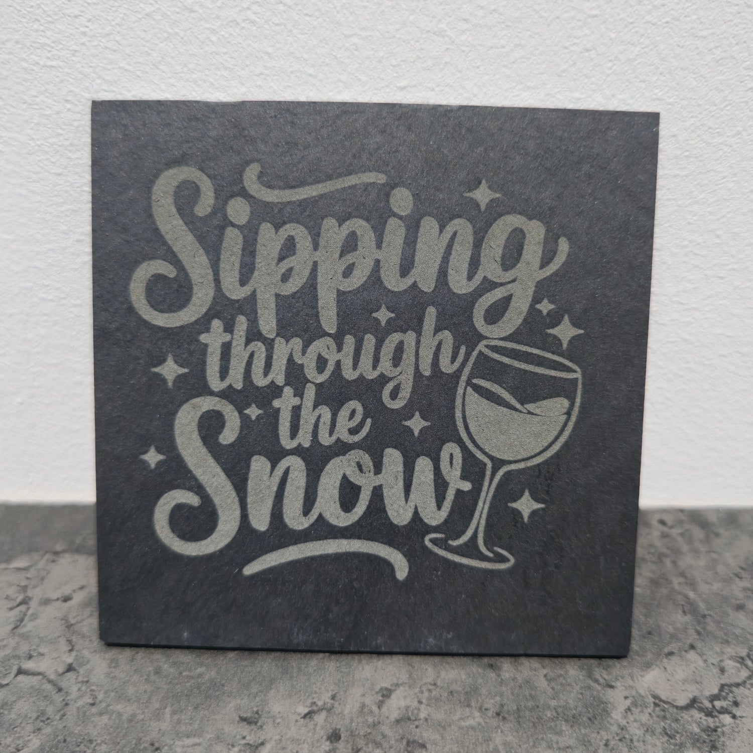 Slate coaster with 'Sipping through the Snow' text and wine glass illustration on a stone surface.
