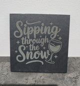 Slate coaster with 'Sipping through the Snow' text and wine glass illustration on a stone surface.