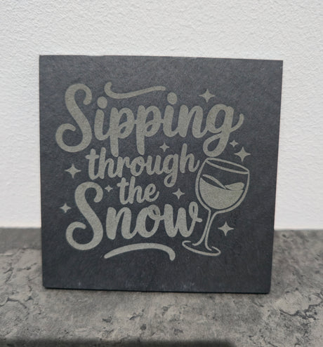 Slate coaster with 'Sipping through the Snow' text and wine glass illustration on a stone surface.