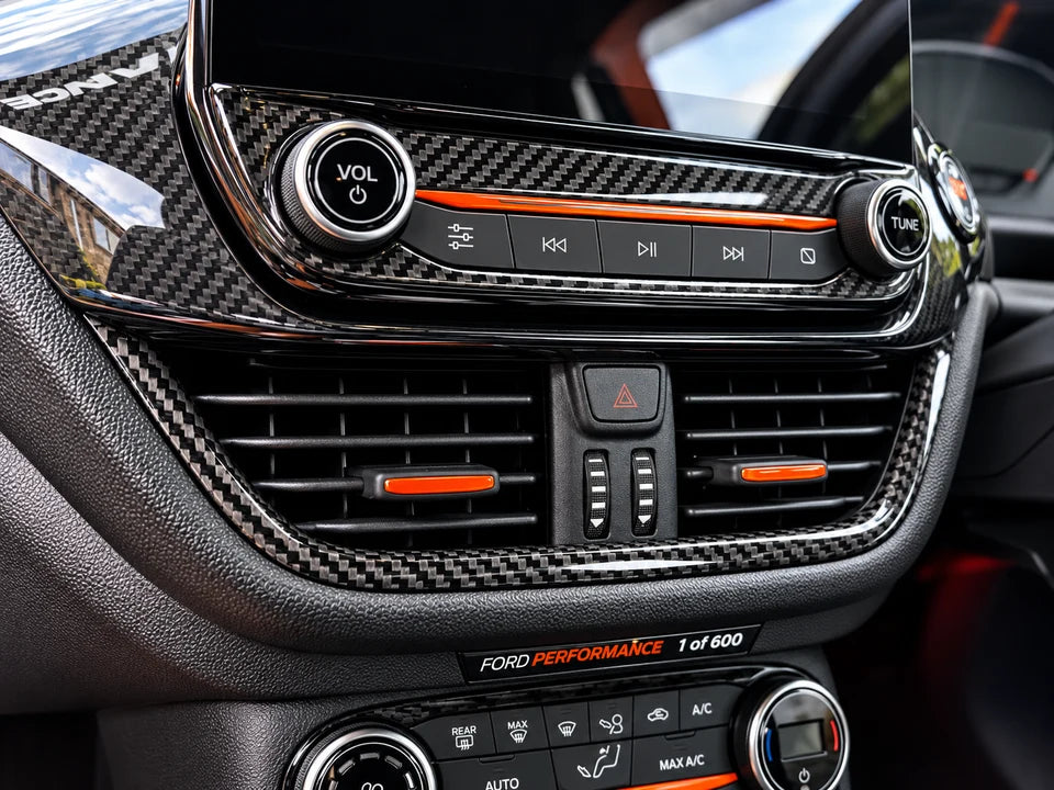 Close-up of a car dashboard with carbon fiber trim, circular control buttons, air vents, and orange accent lines. Visible text includes 'FORD PERFORMANCE 1 of 600'.