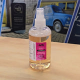 Clear spray bottle with a pink label on a wooden surface, with a blue car and building in the background.