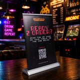 A freestanding black sign on a bar table in a dimly lit pub, with neon signs and an arcade machine blurred in the background. The sign reads “READY 2 ORDER?” with a QR code and small logo text at the top.
