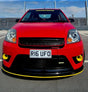 A red sports car parked outdoors on asphalt in front of a fence, with a prominent black front lip spoiler featuring a yellow stripe and matching yellow accents around the fog light openings.
