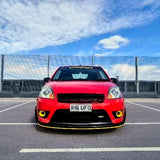 A red hatchback car parked in an outdoor lot under a blue sky with clouds, featuring a black front grille, yellow accents on the bumper, and a license plate reading R16 UFO.