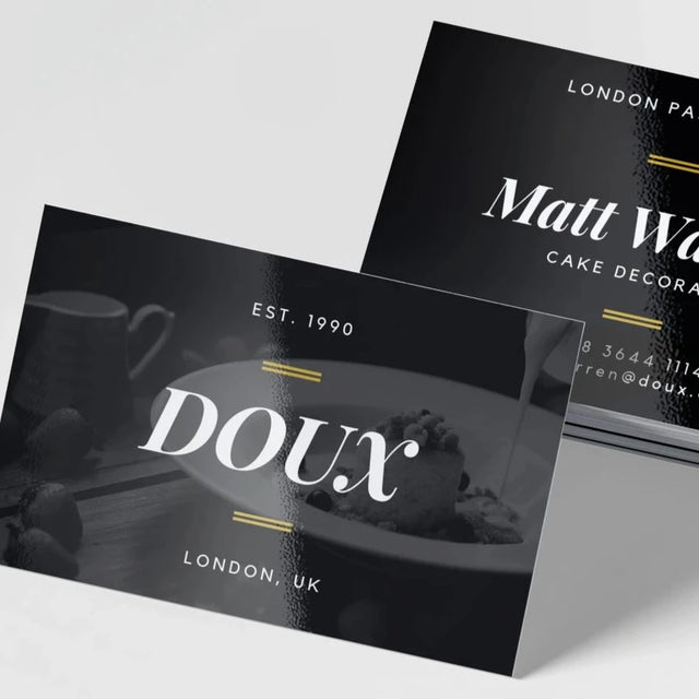 A stack of black glossy business cards with white text and gold accents, featuring the name 'Doux' and 'London UK' on the front.