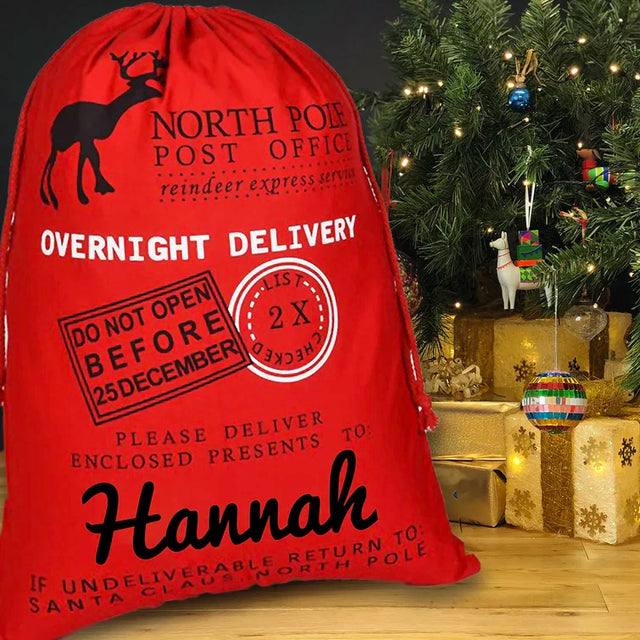 A red gift sack with 'NORTH POLE POST OFFICE' and 'Overnight Delivery' text, along with a name 'Hannah' and a message 'Please do not open before December 25' printed on it, placed near a Christmas tree and gifts.